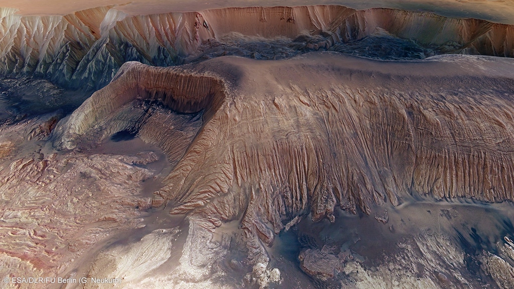 Una vista de la impresionante mesa central dentro de Hebes Chasma. Se ha sacado un trozo en forma de herradura de un lado del montículo (a la izquierda en esta imagen); el material se ha caído al piso del valle a continuación. Un parche oscuro parece acumularse como tinta derramada sobre los restos. Es muy probable que haya material suelto que se haya deslizado por las paredes desde una capa intermedia. El hielo derretido podría haber desempeñado un papel al debilitar las rocas para crear su apariencia fluida. ESA / D